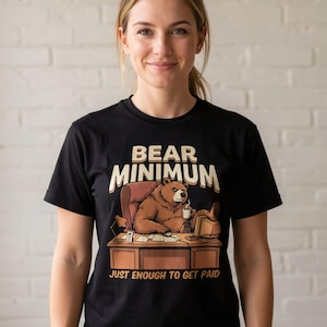 May include: A black short-sleeve t-shirt featuring a graphic of a brown bear sitting at a desk with a computer and coffee mug. The text reads BEAR MINIMUM above the bear and JUST ENOUGH TO GET PAID below the desk in a light tan font color.