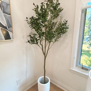 May include: An artificial olive tree in a white pot. The tree has green leaves and small, dark olives. The pot is cylindrical and sits in the corner of a room with light walls and wooden flooring. A framed artwork is visible on the wall.