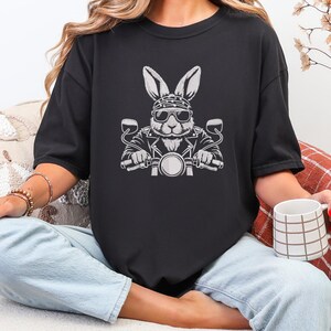 May include: Black t-shirt featuring a white graphic of a rabbit wearing sunglasses and a bandana, riding a motorcycle. The rabbit is depicted with a leather jacket and is holding the handlebars. The design is centered on the front of the shirt.