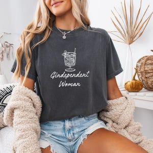 Op de afbeelding: Donkergrijs t-shirt met een cocktail illustratie en de tekst "Gindependent Woman" in het wit. Het shirt heeft een ronde hals en korte mouwen. Het ontwerp toont een cocktail met ijs, een rietje en een garnering.