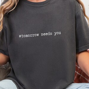 Könnte beinhalten: Dunkelgraues T-Shirt mit dem weißen Text "#tomorrow needs you". Das Shirt hat einen Rundhalsausschnitt und kurze Ärmel. Der Stoff wirkt weich und leicht verwaschen.