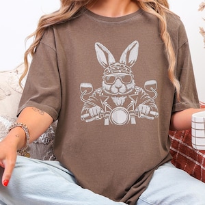 May include: Brown t-shirt featuring a white graphic of a rabbit wearing sunglasses, a bandana, and a leather jacket, riding a motorcycle. The design is centered on the front of the shirt. The shirt has short sleeves and a crew neck.