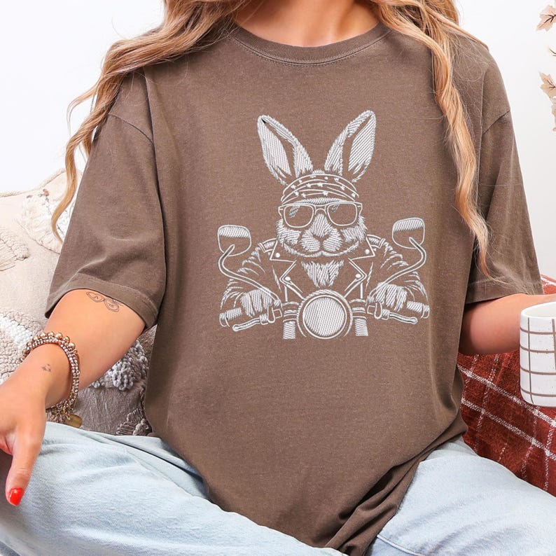 May include: Brown t-shirt featuring a white graphic of a rabbit wearing sunglasses, a bandana, and a leather jacket, riding a motorcycle. The design is centered on the front of the shirt. The shirt has short sleeves and a crew neck.