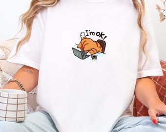 Könnte beinhalten: Weißes T-Shirt mit einem Cartoon-Kater, der seinen Kopf auf einem Laptop ablegt. Der Kater ist orange und braun, mit dem Text "I'm ok!" darüber. Eine kleine Kaffeetasse steht auf dem Schreibtisch. Ein lässiges Kleidungsstück für den Alltag.