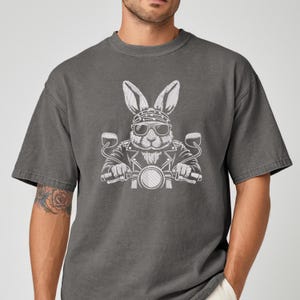 May include: Dark gray t-shirt featuring a white graphic of a rabbit wearing sunglasses and a bandana, riding a motorcycle. The rabbit is depicted in a biker jacket. The shirt has short sleeves and a crew neck.
