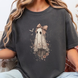May include: Dark gray t-shirt featuring a whimsical ghost design. The ghost is adorned with a bow and floral accents in soft pink and beige tones. Two bats are flying above the ghost. The shirt is a comfortable, casual wear item.
