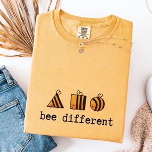 以下が含まれることがあります： マスタードイエローのクルーネックのスウェットシャツ。テキスト「bee different」と蜂をテーマにしたグラフィックが描かれています。繊細な太陽のペンダントが付いたシルバーのネックレスがシャツの上にあります。シャツは、青いジーンズの隣の白い表面に折りたたまれています。