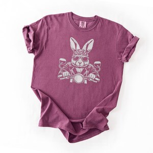 May include: A maroon t-shirt featuring a white graphic of a rabbit wearing sunglasses, a bandana, and a leather jacket, riding a motorcycle. The design is centered on the front of the shirt.