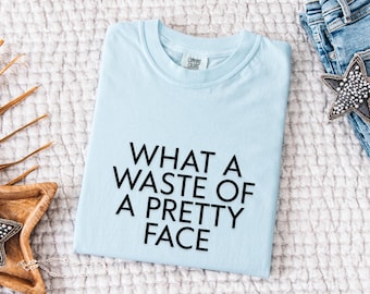 Puede incluir: Camiseta azul claro con la frase negra "WHAT A WASTE OF A PRETTY FACE". La camiseta doblada está sobre una superficie blanca texturizada, con unos vaqueros y un accesorio en forma de estrella en el fondo.