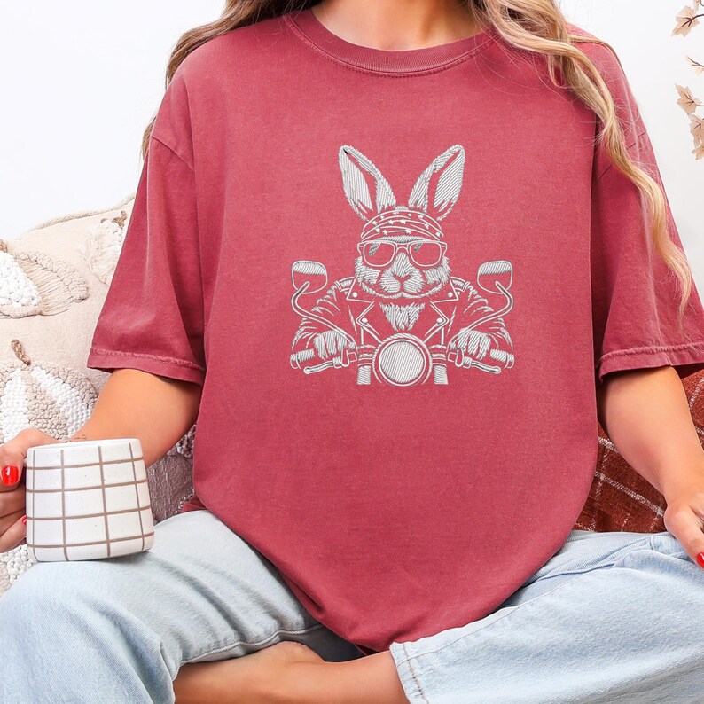 May include: A maroon t-shirt featuring a white graphic of a rabbit wearing sunglasses and a biker jacket, riding a motorcycle. The shirt has short sleeves and a crew neck.
