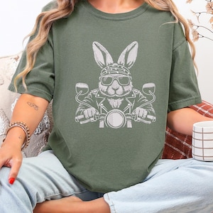 May include: Olive green t-shirt featuring a white graphic of a rabbit wearing sunglasses, a bandana, and a leather jacket, riding a motorcycle. The design is a playful take on biker culture.