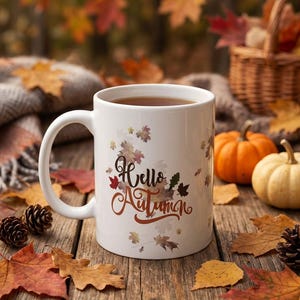 Può includere: Una tazza in ceramica bianca con la scritta "Hello Autumn" in un carattere corsivo marrone, circondata da un motivo stampato di varie foglie autunnali colorate nelle tonalità dell'arancione, del rosso e del marrone, con un manico curvo a sinistra.