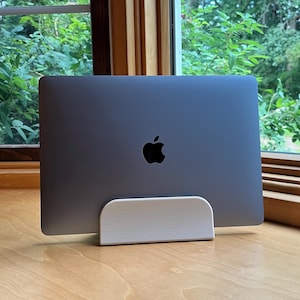 Op de afbeelding: Een zilveren laptop met een zwart Apple-logo staat op een witte houten standaard. De standaard staat op een houten oppervlak, met een raam en groen gebladerte op de achtergrond.