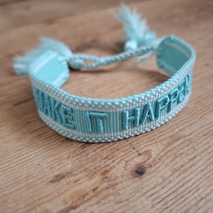 Könnte beinhalten: Ein hellblaues gewebtes Armband mit den Worten "MAKE IT HAPPY", die in einem passenden Blauton gestickt sind. Das Armband hat einen geflochtenen Verschluss mit Quasten. Das Armband liegt auf einer Holzoberfläche.