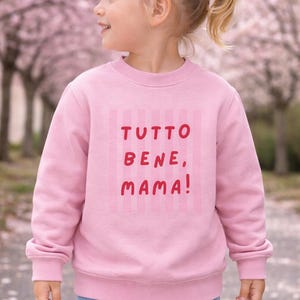 Könnte beinhalten: Ein rosa Sweatshirt mit dem italienischen Satz "TUTTO BENE, MAMA!" in roten Buchstaben. Das Sweatshirt hat einen Rundhalsausschnitt und lange Ärmel. Das Kind trägt eine blaue Jeans.