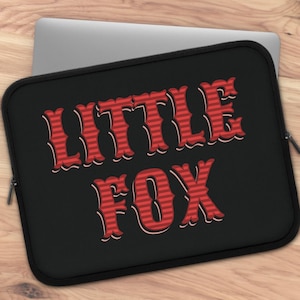 Può includere: Custodia nera per laptop con la scritta "LITTLE FOX" in rosso. La custodia ha una chiusura a zip ed è appoggiata su una superficie di legno. Un computer portatile è parzialmente visibile.