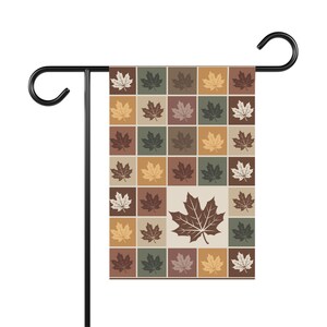 May include: A rectangular garden flag hanging on a black metal stand, featuring a grid pattern of squares in shades of brown, green, and cream. Each square contains a single maple leaf silhouette, with one large central leaf in dark brown.
