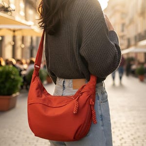 Può includere: Una borsa a tracolla rossa dalla forma curva, con una lunga tracolla regolabile. La borsa ha una chiusura a cerniera e dettagli intrecciati sui lati. La persona indossa un maglione grigio e jeans blu.