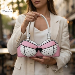 May include: A handbag shaped like a bra with a pink and white polka dot pattern, featuring a small black bow in the center, white trim, and a white top handle. The text "Purseverra" is printed in the upper left corner of the image.
