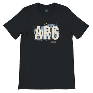 May include: Black t-shirt with the word "ARG" in a cream-colored font, set against a light blue watercolor-style background. Above the text are three gold stars. The text "EST. 1893" is below the main text.