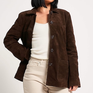 May include: A brown suede jacket featuring a collared neckline, button closure, and two front pockets. The jacket is worn over a white ribbed vest top and light-coloured trousers. The suede has a soft texture, and the style is casual and relaxed.