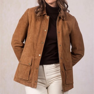 May include: A tan suede jacket with a collared neckline, button closures, and two front pockets. The jacket is worn over a dark brown turtleneck and paired with off-white pants. The jacket has a classic, tailored design.