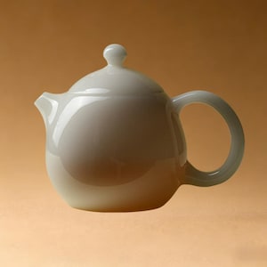 May include: A white ceramic teapot with a rounded body, a curved handle, and a small spout. The lid has a small, round knob. The teapot is set against a warm, neutral background, highlighting its smooth, glossy surface. A classic design for serving tea.