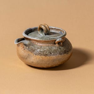 May include: A small, handcrafted ceramic teapot with a lid and spout. The teapot has a rounded body with a textured, earthy appearance, featuring shades of brown, grey, and blue. The lid has a loop handle. The teapot is designed for brewing tea.