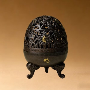 May include: Dark brown, egg-shaped incense burner with intricate floral and dragon cutouts. The burner has a decorative band and sits on three curved feet. A gold-colored accent is visible on the front.