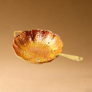 May include: A gold-colored tea strainer with a floral design. The strainer has a handle and a perforated bowl with a textured surface. The color gradient ranges from orange to yellow.