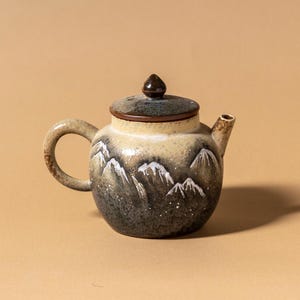 May include: A ceramic teapot with a lid and handle. The teapot is painted with a mountain scene in shades of gray, white, and beige. The lid is dark brown. The teapot is on a beige background.