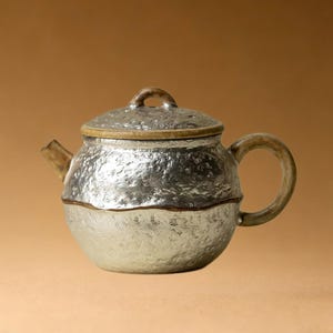 May include: A handcrafted teapot with a textured, metallic silver finish on the upper half and a lighter, matte silver on the lower half. The lid and handle are a contrasting tan color, creating a unique, artisanal look.