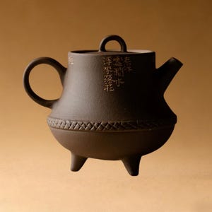 May include: A dark brown ceramic teapot with a round handle and a spout. The teapot has a lid with a small loop handle and three small legs. The body of the teapot has a decorative band around the middle and Chinese characters.