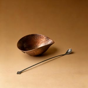 May include: A hammered copper bowl with a unique shape and a long, slender spoon. The bowl has a textured surface and a rich, warm color. The spoon is dark with a decorative end. The items are set against a neutral background.