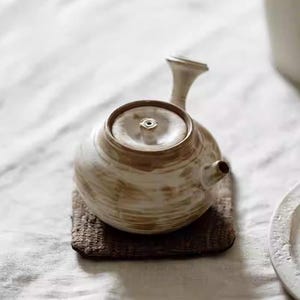 Könnte beinhalten: Eine Keramik-Teekanne mit rundem Körper, Deckel und Ausguss. Die Teekanne ist hellbraun mit dunkleren braunen Streifen und steht auf einem kleinen, quadratischen, dunkelbraunen Untersetzer. Der Griff hat eine schlanke, gebogene Form.