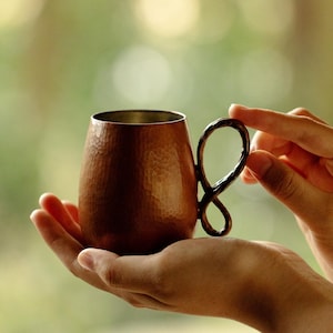 May include: A copper mug with a hammered texture and a unique handle design. The mug is held in two hands against a blurred green background. The interior of the mug appears to be a lighter metal.