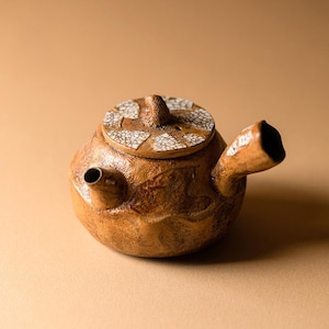 以下が含まれることがあります： 手作りの茶色のセラミックティーポット。表面はざらざらしています。蓋には白と茶色のひび割れ模様があります。注ぎ口とハンドルは丸みを帯びた本体に統合されています。側面に小さな開口部が見えます。