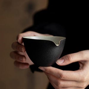 May include: A black ceramic tea cup with a textured exterior and a white interior. The cup has a spout and is held by two hands against a blurred background. The cup is designed for tea ceremonies.
