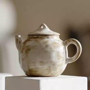 May include: A ceramic teapot with a lid, spout, and handle. The teapot is off-white with brown and tan markings. The teapot is sitting on a white surface.