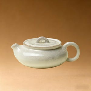 以下が含まれることがあります： 小さなオフホワイトのセラミックティーポット。蓋と丸い取っ手が付いています。 ティーポットには注ぎ口があり、蓋には取り外しを容易にする小さなループが付いています。釉薬はまだら模様です。