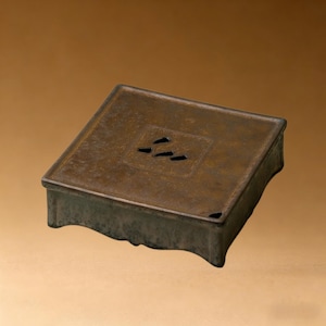 Puede incluir: Un quemador de incienso cuadrado de color bronce con una base ligeramente curvada. La parte superior presenta tres pequeños recortes en forma de onda. El diseño es simple y elegante, adecuado para la decoración del hogar o la meditación.