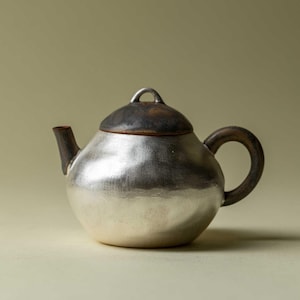 May include: A silver-colored teapot with a dark brown lid and handle. The teapot has a rounded body, a curved spout, and a loop handle. The lid has a small, curved handle. The teapot is likely ceramic or metal.