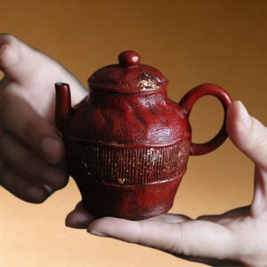 May include: A small, red teapot with a lid and handle. The teapot has a textured surface with vertical lines and gold accents. The teapot is held in two hands against a warm, neutral background.