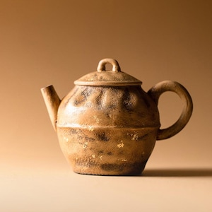 May include: A handcrafted, brown ceramic teapot with a lid and handle. The teapot has a textured surface with gold accents. The spout and handle are curved. The teapot is designed for brewing tea.