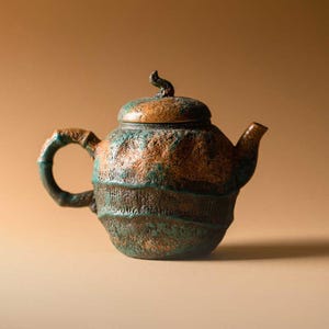 May include: A textured ceramic teapot with a lid and curved spout. The teapot is finished in shades of brown, green, and copper, giving it an aged appearance. The handle is a curved loop. A small handle sits atop the lid.