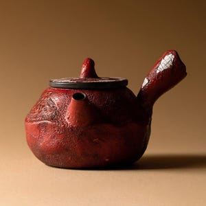 May include: A red ceramic teapot with a textured surface. The teapot has a small spout, a handle with white accents, and a lid with a black rim. The teapot is designed for brewing tea.