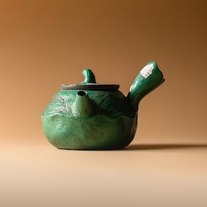 Może przedstawiać: Zielony ceramiczny czajnik o teksturowanej powierzchni. Czajnik posiada dzióbek, uchwyt i pokrywkę. Uchwyt ma biało-szary wzór. Czajnik stoi na beżowym tle.