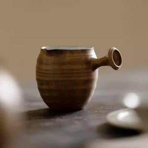 Könnte beinhalten: Eine handgefertigte, braune Keramik-Tasse mit einem einzigartigen Griffdesign. Die Tasse hat einen abgerundeten Körper mit horizontalen Rillen und einen kleinen, kreisförmigen Griff. Der Innenrand ist heller, und die Tasse scheint aus Steinzeug zu sein.