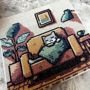 May include: Cross-stitch art piece depicting a cat relaxing on a couch. The artwork features a cozy living room scene with a plant, picture frame, and coffee cup. The color palette includes shades of beige, brown, and blue.