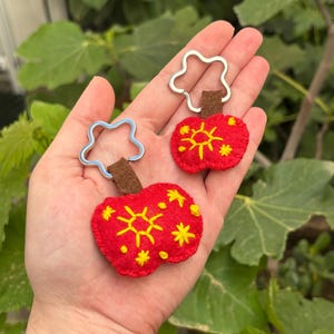 Puede incluir: Llaveros artesanales de fieltro rojo con diseños de sol y estrellas bordados en amarillo. Cada llavero tiene un tallo de fieltro marrón y un anillo de metal plateado. Los llaveros se sostienen en una mano.